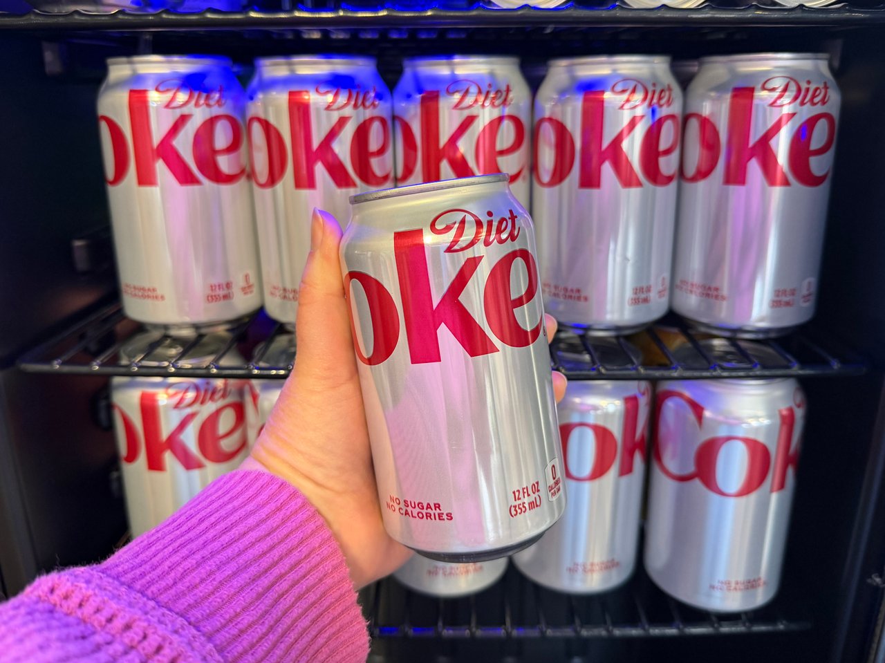 Person holding a Diet Coke in front of a refrigerator full of them.