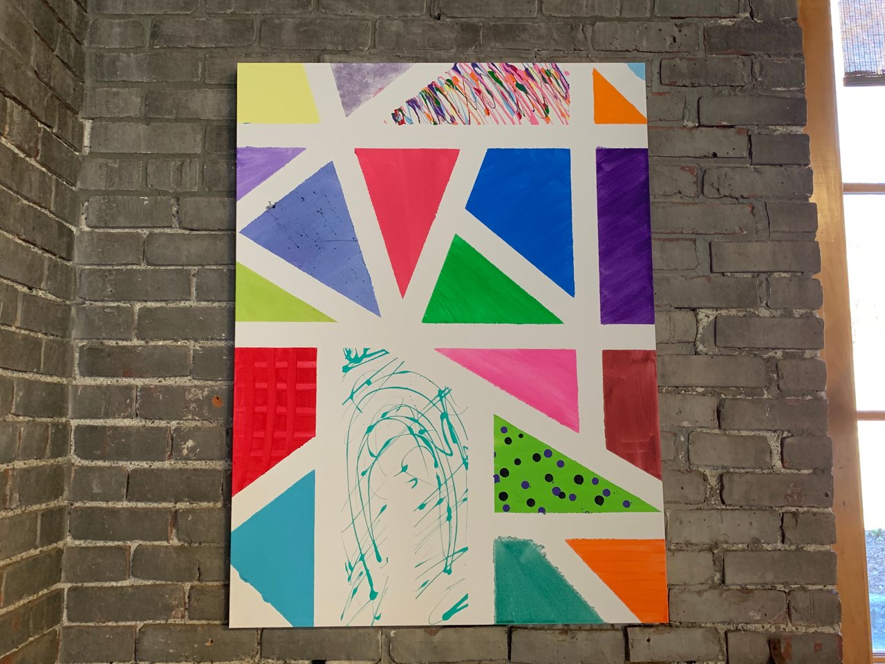 An abstract, colorful painting against a gray brick wall.