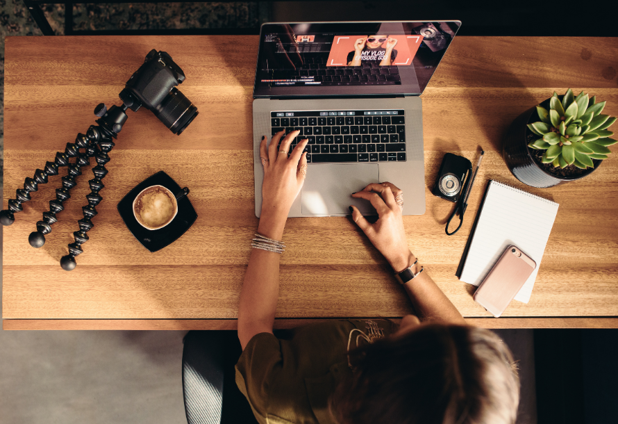 An overhead shot of someone working on a laptop, with a camera on a tripod and a coffee.